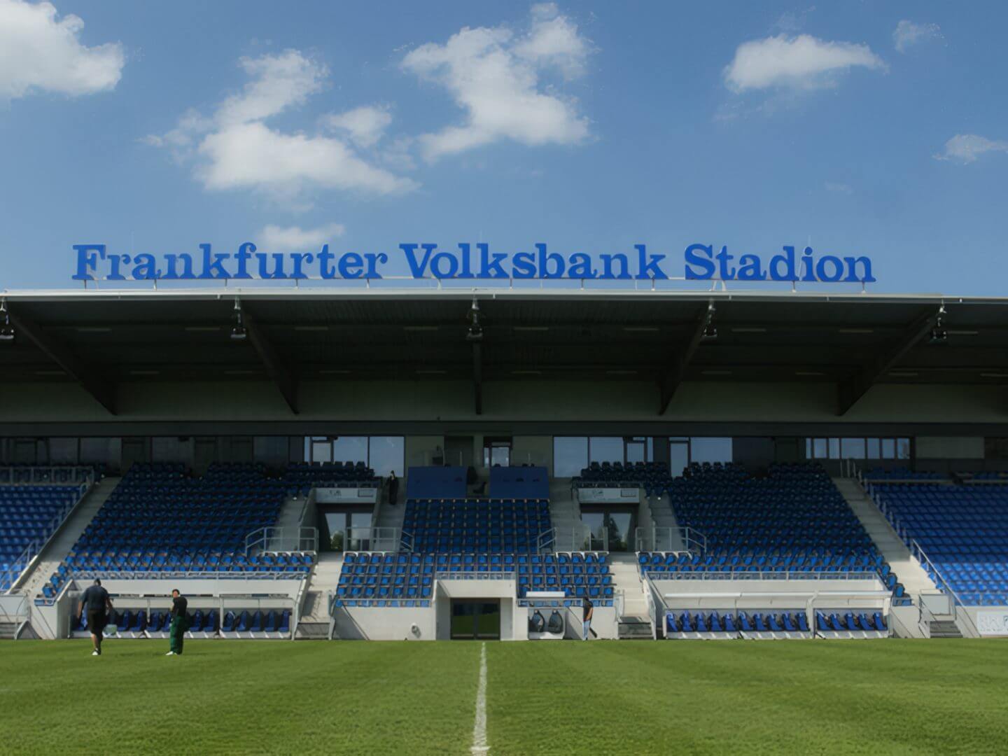 Ansicht der BBBank Arena in Frankfurt mit leeren blauen Sitzreihen und grünem Spielfeld unter blauem Himmel.