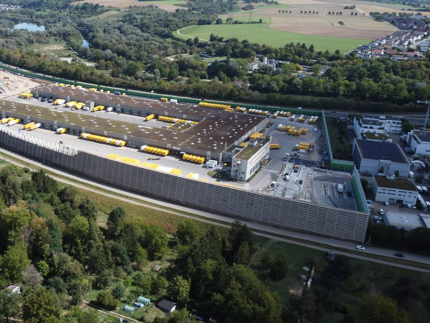 Luftaufnahme des DHL Logistikzentrums in Köngen, realisiert von der Hellmich Gruppe, mit zahlreichen gelben Transportfahrzeugen vor dem Gebäude. Luftaufnahme des DHL Logistikzentrums in Köngen, realisiert von der Hellmich Gruppe, mit zahlreichen gelben Transportfahrzeugen vor dem Gebäude.
