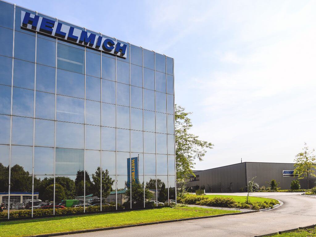 Moderne Firmenzentrale von Hellmich mit spiegelnder Glasfassade und grünem Vorgarten unter blauem Himmel.