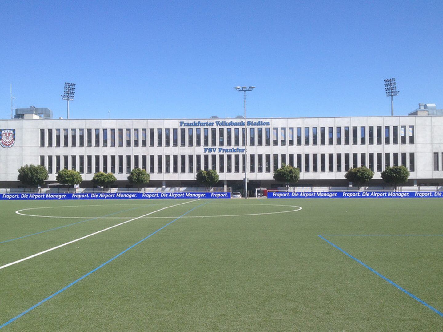 Das Frankfurter Volksbank Stadion mit klarer Fassade und Sportplatz, ein Bauprojekt der Hellmich Gruppe, präsentiert sich an einem sonnigen Tag unter blauem Himmel.