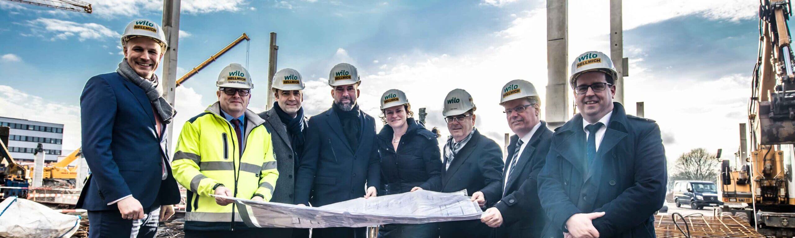 Fünf Teammitglieder mit HELLMICH-Schutzhelmen schauen gemeinsam auf Baupläne unter freiem Himmel.