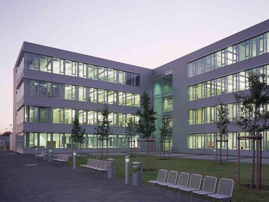 Beleuchtetes Bürogebäude mit Sitzgelegenheiten und Bäumen im Gewerbepark Kamp-Lintfort bei Abenddämmerung.