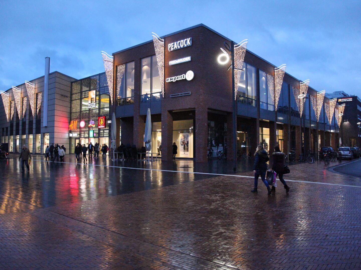 Abendaufnahme der Neutor Galerie in Dinslaken, ein modernes Bauprojekt der Hellmich Gruppe mit beleuchteten Fassaden und Passanten auf dem Platz davor. Abendaufnahme der Neutor Galerie in Dinslaken, ein modernes Bauprojekt der Hellmich Gruppe mit beleuchteten Fassaden und Passanten auf dem Platz davor.